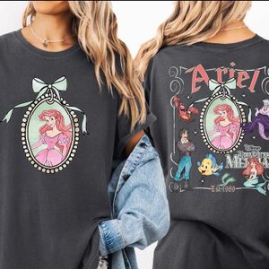 two t-shirts with the character Ariel from the Disney animated film "The Little Mermaid". The t-shirt on the left has a portrait of Ariel and the text "Ariel" above her, while the t-shirt on the right has a graphic of Ariel surrounded by various sea creatures and the text "Ariel" and "The Little Mermaid" on the back.