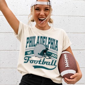 Może przedstawiać: Kremowy t-shirt z kr&oacute;tkim rękawem z nadrukiem graficznym zawierającym napis PHILADELPHIA, EST. 1933 oraz Football. Wz&oacute;r zawiera ilustrację szarego kasku do futbolu amerykańskiego w odcieniach ciemnego turkusu i szarości oraz brązową piłkę.
