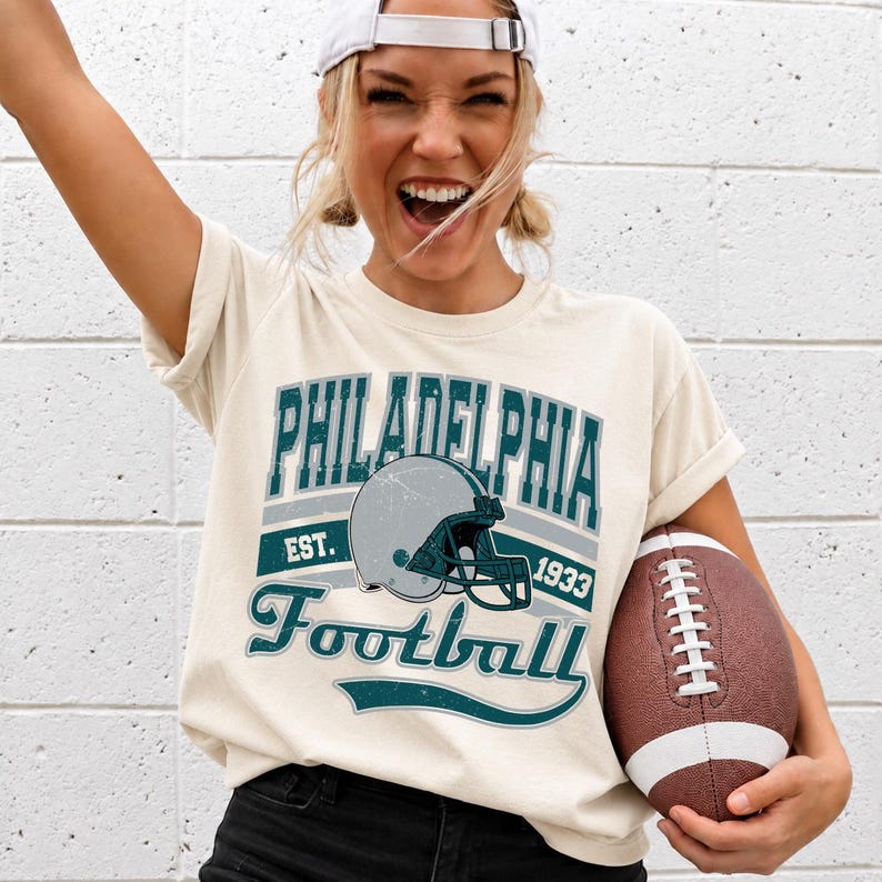 Może przedstawiać: Kremowy t-shirt z kr&oacute;tkim rękawem z nadrukiem graficznym zawierającym napis PHILADELPHIA, EST. 1933 oraz Football. Wz&oacute;r zawiera ilustrację szarego kasku do futbolu amerykańskiego w odcieniach ciemnego turkusu i szarości oraz brązową piłkę.