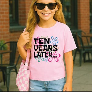 May include: A light pink t-shirt with the text "TEN YEARS LATER..." in black, with colorful flower designs. The shirt is worn by a person wearing sunglasses and carrying a pink and white checkered tote bag.