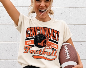 Puede incluir: Una camiseta de manga corta color crema con un gráfico desgastado que incluye el texto CINCINNATI, EST. 1968 y Football. El diseño presenta un casco de fútbol americano negro con una máscara naranja, rayas naranjas y negras y letras.
