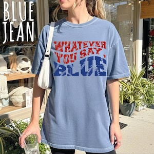 May include: A blue jean colored short-sleeve t-shirt featuring the red and blue text "WHATEVER YOU SAY, BLUE" with a baseball stitch pattern integrated into the letters. The shirt has a crew neckline and a relaxed fit, worn with a white shoulder bag.