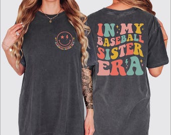 May include: Dark gray t-shirt with the text "IN MY BASEBALL SISTER ERA" on the back in retro colors. The front features a smiley face with lightning bolts and the words "BASEBALL SISTER ERA".