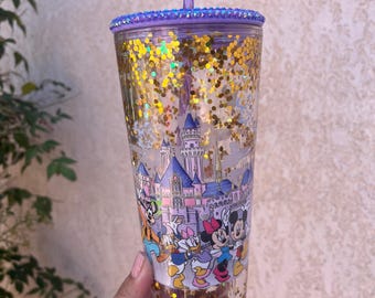 May include: A clear plastic tumbler with a purple glitter rim and a purple straw. The tumbler is filled with gold glitter and features a cartoon image of Mickey Mouse, Minnie Mouse, Donald Duck, and Goofy in front of a pink castle.