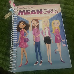 May include: A spiral-bound notebook featuring a Mean Girls Little Golden Book cover illustration of four characters in a school hallway. The cover displays the title Mean Girls in large purple text and includes decorative ribbons attached to the white coil.