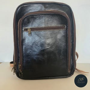 Könnte beinhalten: Ein schwarzer Lederrucksack mit braunen Verzierungen und einer Reißverschlusstasche auf der Vorderseite. Der Rucksack hat einen Tragegriff oben und verstellbare Träger.