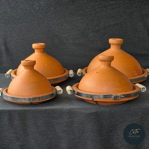 Può includere: Quattro tagine in terracotta con manici in legno e bordi in metallo. I tagine sono disposti in fila su una superficie grigio scuro.
