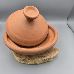 Può includere: Una pentola tagine in terracotta con coperchio conico e base rotonda. La pentola è realizzata in argilla non smaltata e poggia su una piccola lastra di legno. Il tagine è un tradizionale recipiente di cottura nordafricano.