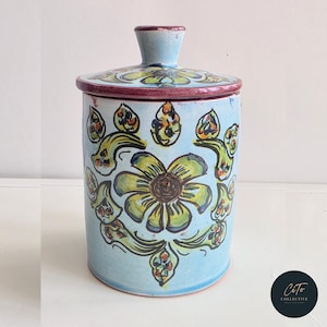 Può includere: Un barattolo di ceramica azzurra con coperchio, con un motivo floreale dipinto a mano in giallo, verde e marrone. Il barattolo ha una forma cilindrica con coperchio e una piccola maniglia. Il bordo del coperchio è rosso scuro.