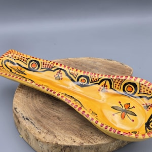 Könnte beinhalten: Ein gelber Keramik-Löffelhalter mit einem schwarzen und roten geometrischen Muster. Der Löffelhalter ist mit einer stilisierten Blume und Punkten verziert.