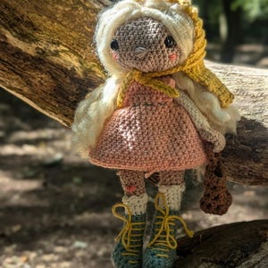 May include: A crocheted doll with a pink dress, yellow and white hooded jacket, and brown boots. The doll is sitting on a tree trunk in a forest setting.
