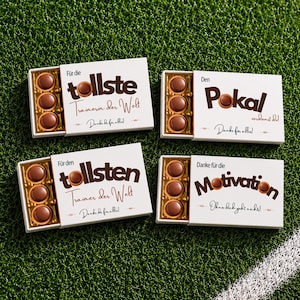 Könnte beinhalten: Vier rechteckige weiße Geschenkboxen mit jeweils drei runden Milchschokoladen. Die Boxen sind mit braunem Text auf Deutsch bedruckt, darunter Phrasen wie 'tollste Trainerin der Welt', 'Pokal', 'tollsten Trainer der Welt' und 'Motivation'.