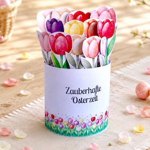 Può includere: Un contenitore cilindrico bianco con tulipani di carta colorati, il testo "Zauberhafte Osterzeit" e un bordo floreale. Petali sparsi e uova decorate sono su un tavolo con un cestino di uova e fiori.