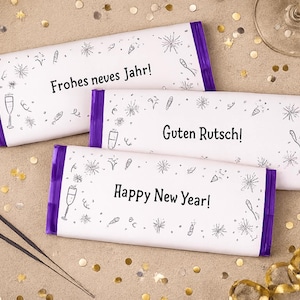 Peut inclure: Trois tablettes de chocolat rectangulaires emballées dans du papier blanc avec des extrémités violettes. Les emballages présentent des dessins au trait noir de feux d'artifice et de flûtes à champagne. Le texte indique "Frohes neves Jahr!", "Guten Rutsch!" et "Happy New Year!" en noir.