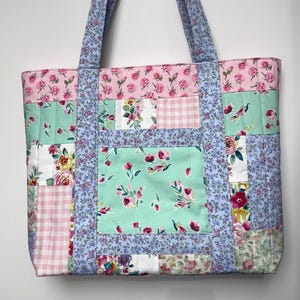 Puede incluir: Un bolso tote patchwork con estampados florales y de cuadros en rosa, verde y blanco. El bolso tiene asas y bordes florales de color azul claro. El bolso está hecho de tela acolchada.
