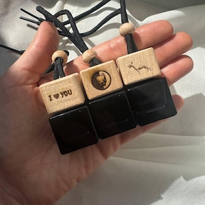 May include: Three wooden car air fresheners with black glass bottles. The wooden blocks have different designs, including a heart with the text "I LOVE YOU", a geometric design, and a stylized animal.