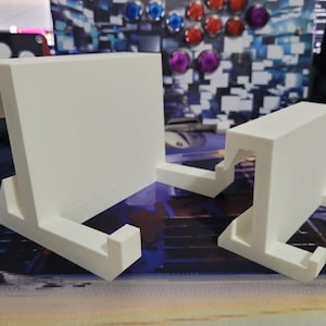May include: Two white plastic phone stands, one larger than the other, with a unique design featuring a curved lip on the top and a base with two angled supports. The stands are on a patterned surface.