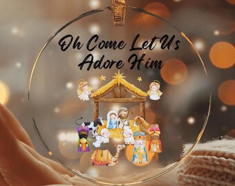 Puede incluir: Un adorno navideño de vidrio transparente con una estrella dorada y las palabras "Oh Come Let Us Adore Him" impresas en él. El adorno presenta una representación de la escena de la natividad con el niño Jesús, María, José y los tres reyes magos.