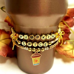 May include: Three beaded bracelets with the words "PUMPKIN SPICE" spelled out in gold letter beads. A charm of a coffee cup with a pumpkin is attached to the bottom bracelet. The bracelets are wrapped around a brown tumbler.