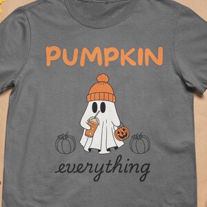May include: A gray t-shirt with the words "PUMPKIN everything" in orange and black. The shirt features a cartoon ghost wearing an orange beanie and holding a pumpkin spice drink and a pumpkin. Two pumpkins are on either side of the ghost.
