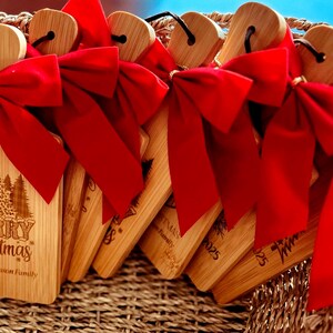 May include: A collection of wooden Christmas ornaments with red bows. Each ornament has a heart-shaped top and a rectangular body with the words "Merry Christmas" and a forest design engraved on it. The ornaments are in a woven basket.