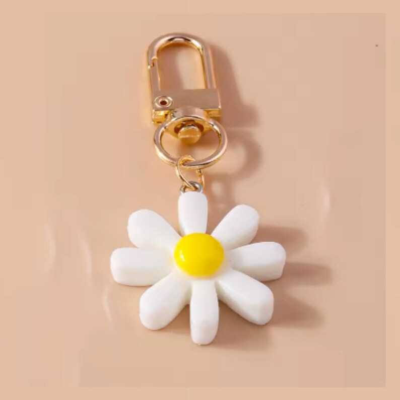 May include: A gold-toned keychain with a white daisy charm. The daisy has a yellow center and white petals.