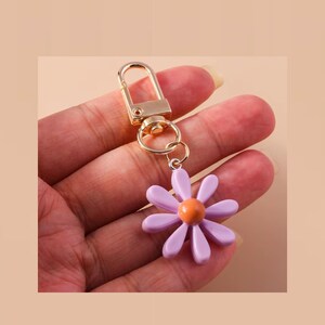 May include: A gold key chain with a purple flower charm. The flower has a light orange center.