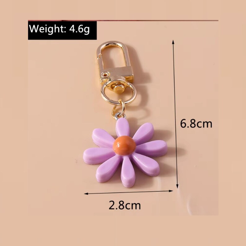 May include: A purple flower keychain with a gold metal clasp and ring. The flower has an orange center and measures 2.8 inches in diameter. The keychain is 6.8 inches long and weighs 4.6 grams.