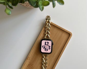 以下が含まれることがあります： ヘリンボーン柄のゴールドの腕時計バンド。時計の文字盤は黒で、ピンクの背景に白い数字があります。時計は木製の表面にあります。
