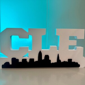 May include: White wooden sign with the letters "CLE" in a bold, blocky font. The letters are cut out to reveal a black silhouette of the Cleveland skyline.