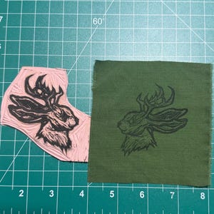 Op de afbeelding: Een roze rubberen linosnijblok met een uitgesneden jackalope-ontwerp naast een vierkante olijfgroene stoffen lap met dezelfde jackalope-afbeelding in zwarte inkt, beide rustend op een groene snijmat met witte rasterlijnen en maataanduidingen.