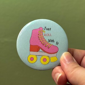 May include: A round, light blue button with a pink roller skate illustration and the text "Just Roll With It". The roller skate has yellow laces and wheels, and an orange heel.