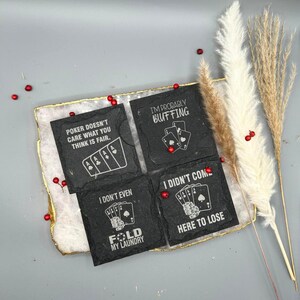 May include: Four black slate coasters with poker-themed quotes and card illustrations. The coasters are arranged on a decorative tray with a gold edge. The quotes include "Poker doesn't care what you think is fair" and "I'm probably bluffing."