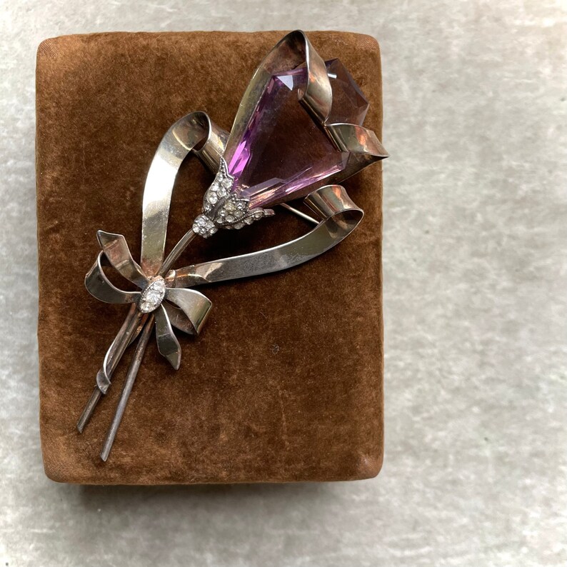 May include: A vintage silver-tone brooch in the shape of a flower with a large, faceted purple gemstone center. The brooch features a ribbon design and is embellished with small, clear crystals. The brooch is displayed on a brown velvet surface.