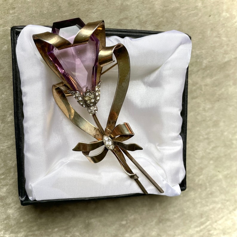 May include: A vintage brooch in the shape of a flower, presented in a black box with white satin. The brooch features a large, faceted, light purple gemstone as the flower's head, with silver-toned metal ribbons and a bow. Small clear crystals accent the flower's base and bow.