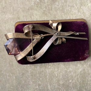 May include: A vintage silver-tone brooch featuring a large, faceted, light purple gemstone shaped like a rose. The brooch has a ribbon and bow design, set against a dark purple velvet background.