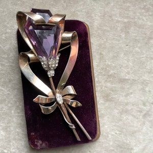 May include: A vintage brooch in the shape of a flower bouquet. The brooch features a large, faceted purple gemstone at the top, with a silver-toned ribbon and stem design. The flower's stem is adorned with small, clear gemstones. The brooch is set against a dark purple velvet background.