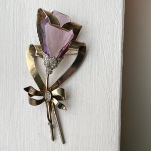 May include: A vintage brooch in the shape of a flower. The brooch features a faceted, light purple crystal flower head, set in a gold-toned metal frame with a ribbon and bow design. The flower's stem is adorned with small, clear crystals.