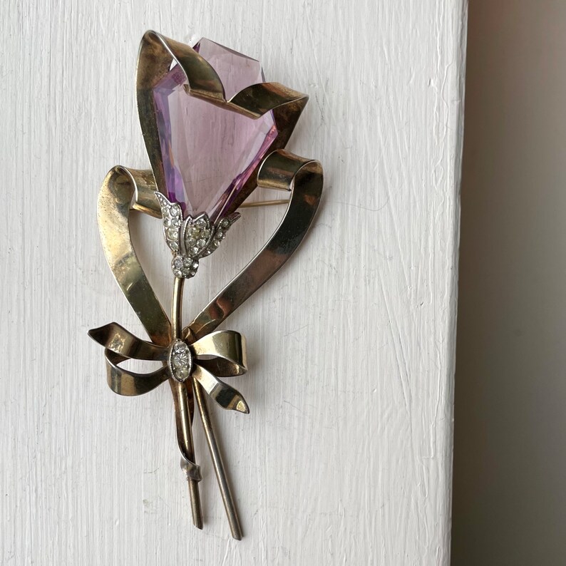 May include: A vintage brooch in the shape of a flower. The brooch features a faceted, light purple crystal flower head, set in a gold-toned metal frame with a ribbon and bow design. The flower's stem is adorned with small, clear crystals.