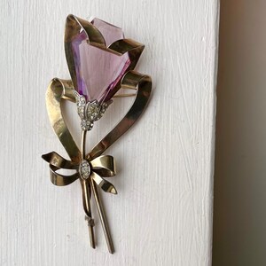 May include: A vintage brooch in the shape of a flower. The flower has a faceted, light purple crystal bloom, with a stem and ribbon made of gold-toned metal. The flower's center and the ribbon's knot are adorned with small, clear crystals.
