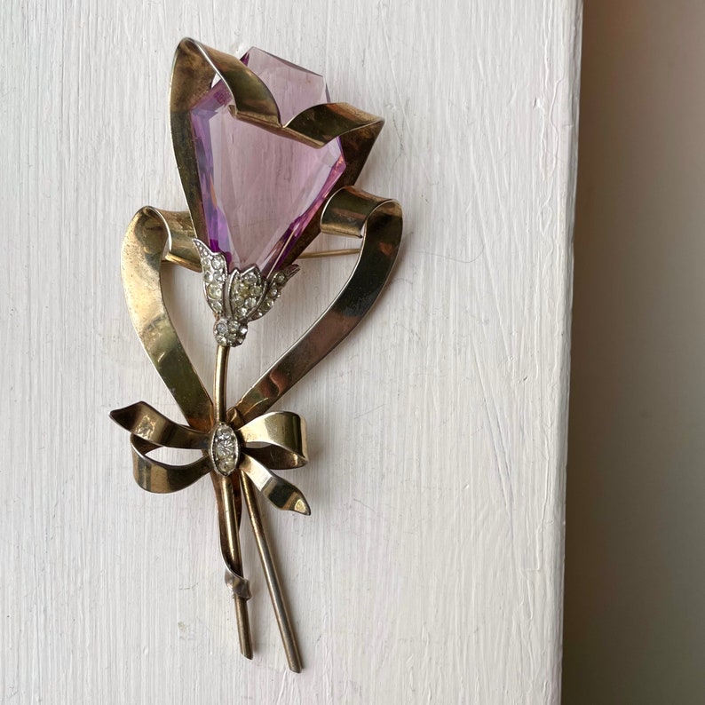 May include: A vintage brooch in the shape of a flower. The flower has a faceted, light purple crystal bloom, with a stem and ribbon made of gold-toned metal. The flower's center and the ribbon's knot are adorned with small, clear crystals.