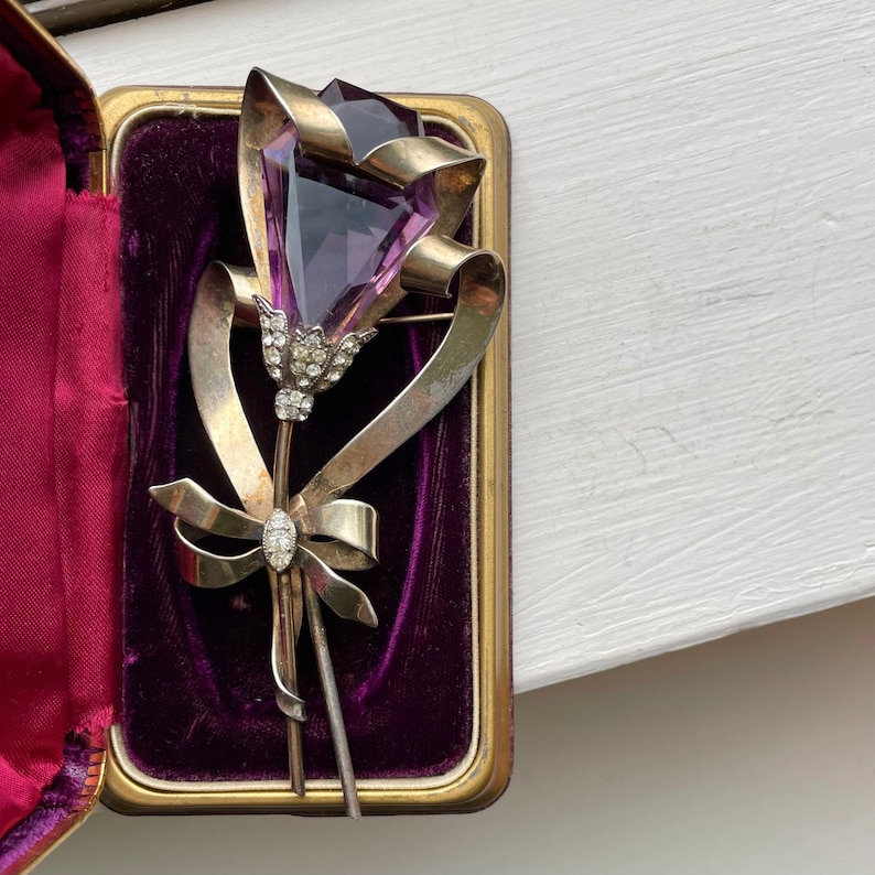 May include: Vintage floral brooch featuring a large, faceted purple gemstone as the flower's head. The brooch has a ribbon design with a bow and is set with small, clear gemstones. The brooch is displayed in a gold-framed case with a purple velvet interior.