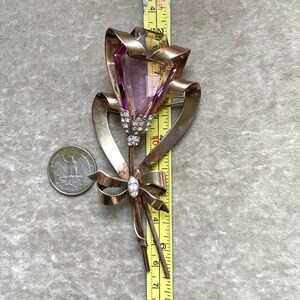 May include: A vintage silver-toned brooch in the shape of a flower, featuring a large, faceted purple gemstone. The brooch includes a ribbon design and is accented with small, clear crystals. The brooch measures approximately 3 inches in length.