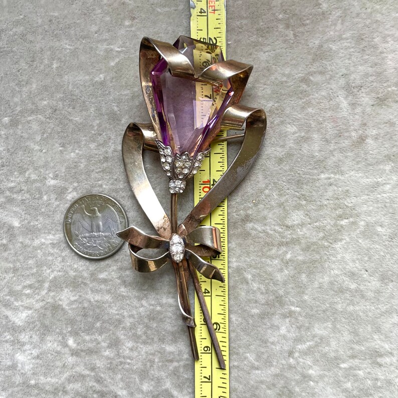 May include: A vintage silver-toned brooch in the shape of a flower, featuring a large, faceted purple gemstone. The brooch includes a ribbon design and is accented with small, clear crystals. The brooch measures approximately 3 inches in length.