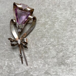 May include: A vintage silver-toned brooch in the shape of a flower. The flower features a large, faceted, purple gemstone as the bloom, with a ribbon and bow design. The brooch is embellished with small, clear gemstones.