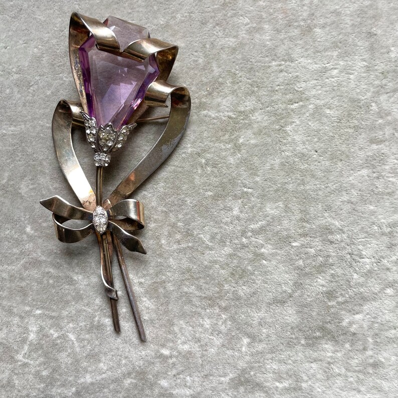 May include: A vintage silver-toned brooch in the shape of a flower. The flower features a large, faceted, purple gemstone as the bloom, with a ribbon and bow design. The brooch is embellished with small, clear gemstones.