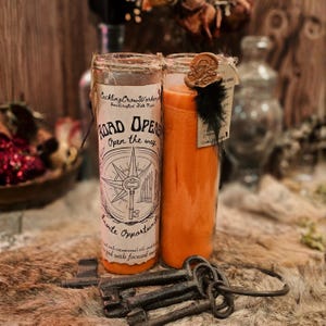 May include: Two orange candles in glass jars with the text "Road Opener" and a compass design. A bundle of antique keys and a black feather are also visible. The candles are on a fur-like surface.