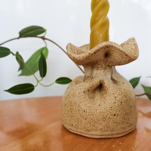 Peut inclure: Un bougeoir en céramique en forme de sac avec un haut à volants. Le bougeoir est moucheté de brun et de beige et contient une bougie jaune.