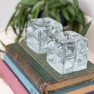 Puede incluir: Dos candelabros cuadrados de vidrio transparente con un diseño geométrico texturizado se encuentran sobre una pila de libros antiguos. Los libros están encuadernados en varios colores, incluyendo verde, azul y rosa. Una planta verde está en el fondo.