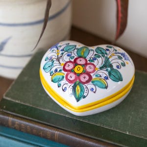 May include: A heart-shaped, white ceramic trinket box with a hand-painted floral design. The lid features a central pink flower with a yellow center, surrounded by green leaves and blue accents. The box has a yellow rim and sits atop a stack of books.